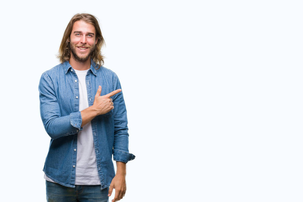 Young handsome man with long hair over isolated background cheerful with a smile of face pointing with hand and finger up to the side with happy and natural expression on face looking at the camera. - Photo, Image