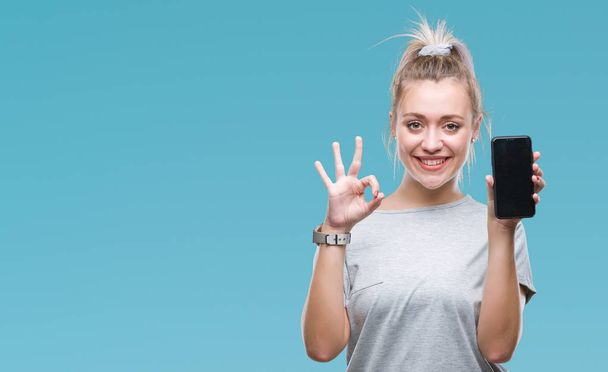 Young blonde woman showing screen of smartphone over isolated background doing ok sign with fingers, excellent symbol - Foto, imagen