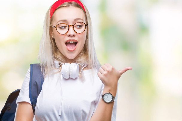 Jonge blonde student vrouw het dragen van bril en rugzak op geïsoleerde achtergrond glimlachend met blij gezicht kijken en wijzen naar de kant met duim omhoog. - Foto, afbeelding