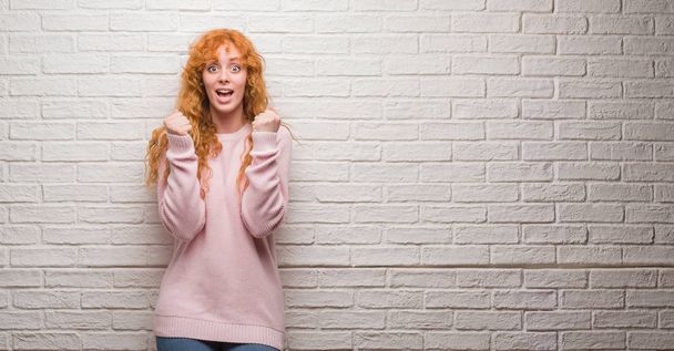 Young redhead woman standing over brick wall celebrating surprised and amazed for success with arms raised and open eyes. Winner concept. - Foto, afbeelding
