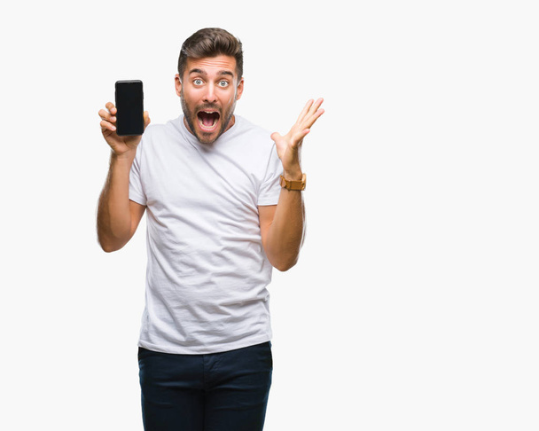 Young handsome man showing smartphone screen over isolated background very happy and excited, winner expression celebrating victory screaming with big smile and raised hands - Photo, Image