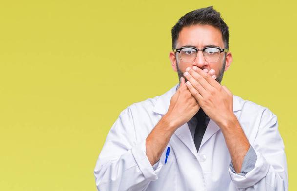 Erwachsener hispanischer Wissenschaftler oder Arzt, der weißen Mantel über isoliertem Hintergrund trägt, schockiert, den Mund mit den Händen versehentlich zu bedecken. Geheimes Konzept. - Foto, Bild