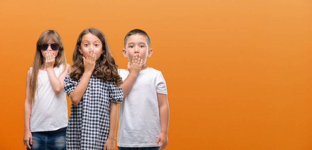Группа мальчишек и девчонок за оранжевым фоном прикрывают рот рукой, потрясенной стыдом за ошибку, выражением страха, испуганной в тишине, секретной концепцией
 - Фото, изображение