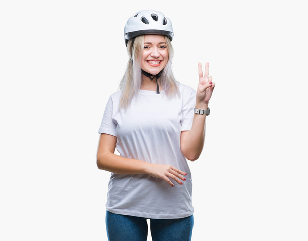 Jonge blonde vrouw fietser veiligheid helm dragen over geïsoleerde achtergrond glimlachend met blij gezicht winking op de camera doen overwinning teken. Nummer twee. - Foto, afbeelding