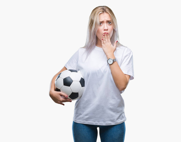 Молодая блондинка, держащая футбольный мяч на изолированном заднем плане, прикрывает рот рукой, потрясенной стыдом за ошибку, выражением страха, испуганной в тишине, секретной концепцией
 - Фото, изображение