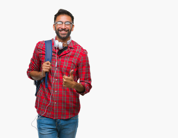 Σπουδαστής ενηλίκων Ισπανόφωνος άνθρωπος φορώντας ακουστικά και σακίδιο πάνω από απομονωμένες φόντο κάνει ευτυχισμένη μπράβο χειρονομία με το χέρι. Την έγκριση έκφραση κοιτάζοντας την κάμερα με δείχνει επιτυχία. - Φωτογραφία, εικόνα