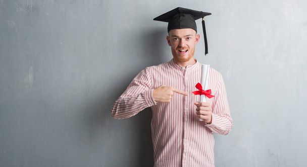 Jovem ruiva homem sobre parede grunge cinza vestindo graduação cap segurando grau muito feliz apontando com a mão e o dedo
 - Foto, Imagem