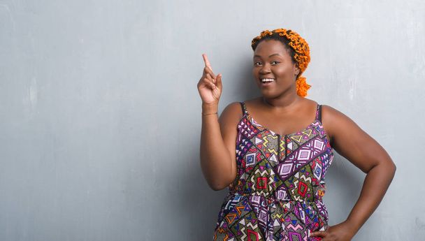 Nuori afrikkalainen amerikkalainen nainen yli harmaa grunge seinä yllään oranssi punokset sormella ylös onnistunut idea
 - Valokuva, kuva