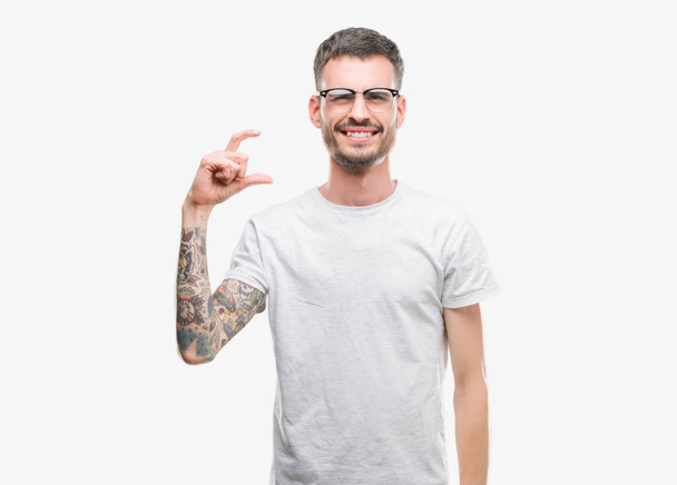Jeune homme adulte tatoué sourire et geste confiant avec la main faisant signe de taille avec les doigts tout en regardant et la caméra. Concept de mesure
. - Photo, image