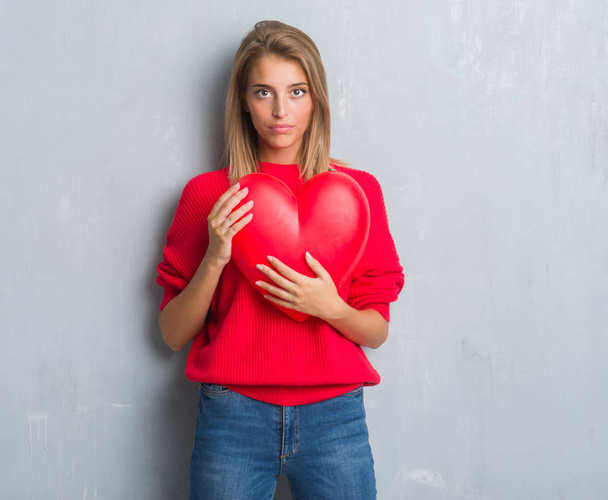Mooie jonge vrouw over grunge grijs muur holding rood hart met een vertrouwen uitdrukking op slimme gezicht ernstige denken - Foto, afbeelding