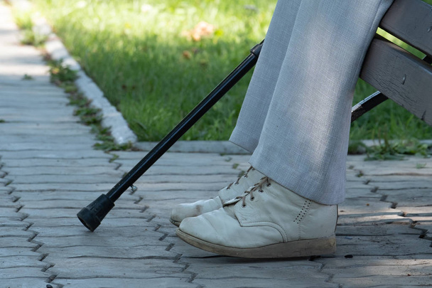 En la imagen de un pie, una muleta y un banco en el parque. En la imagen de una pierna en pantalones viejos, calzados en zapatos viejos, una muleta y un banco en el parque
. - Foto, Imagen