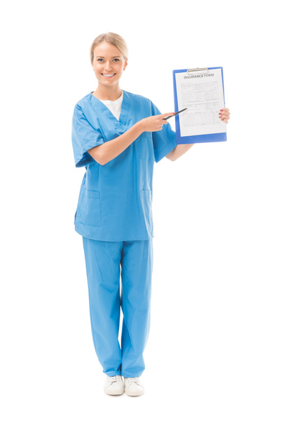 lachende jonge verpleegster wijzend op Klembord met verzekeringsformulier geïsoleerd op wit - Foto, afbeelding