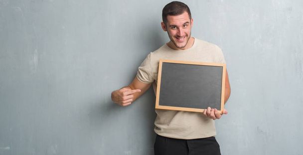 Young caucasian man over grey grunge wall holding blackboard very happy pointing with hand and finger - Photo, Image