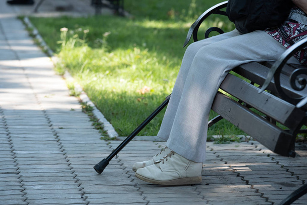 On the image of a foot, a crutch and a bench in the park. On the image of a leg in old pants, shod in old shoes, a crutch and a bench in the park. - Photo, Image