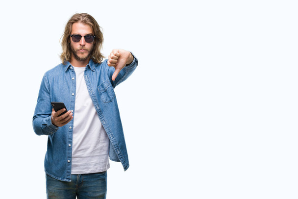 Young handsome man with long hair over isolated background sending message using smartphone with angry face, negative sign showing dislike with thumbs down, rejection concept - Foto, Imagen