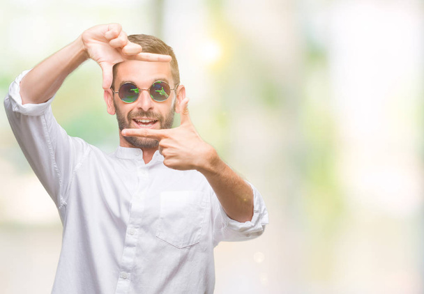 Young handsome man wearing glasses over isolated background smiling making frame with hands and fingers with happy face. Creativity and photography concept. - Photo, Image