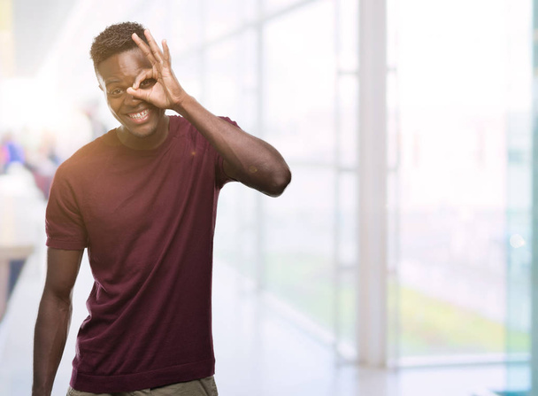 Jonge Afro-Amerikaanse man met blij gezicht lachend doen ok teken met de hand op oog kijken door de vingers - Foto, afbeelding