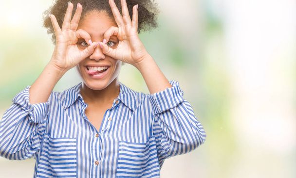 Young afro american woman over isolated background doing ok gesture like binoculars sticking tongue out, eyes looking through fingers. Crazy expression. - Foto, immagini