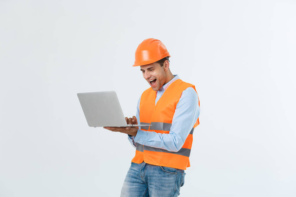 Volwassen man constructor verrast en werken met de laptop in de helm binnenshuis. - Foto, afbeelding