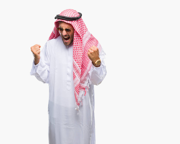 Jovem homem bonito vestindo keffiyeh sobre fundo isolado muito feliz e animado fazendo gesto vencedor com os braços levantados, sorrindo e gritando por sucesso. Conceito de celebração
. - Foto, Imagem