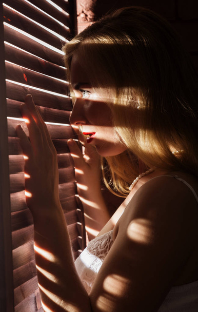 Kaunis nainen alusvaatteet stending lähellä ikkunaa ja suljettu louver aurinkoisena päivänä. Raidallinen varjo vartalossa - Valokuva, kuva