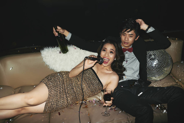 Παρέα στο νυχτερινό κέντρο διασκέδασης: όμορφος ασιατικό άτομο με μπουκάλι κρασί κάθεται στον άνετο καναπέ και να βλέπουν φωτογραφική μηχανή, ενώ το karaoke τραγούδι ελκυστική νεαρή φιλενάδα - Φωτογραφία, εικόνα