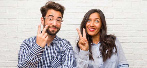 Young indian woman and caucasian man couple showing number two, symbol of counting, concept of mathematics, confident and cheerful - Foto, Imagen
