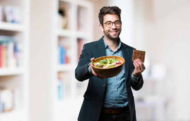 young man choosing between chocolate and salad on blurred background - Photo, Image