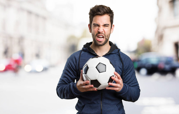 joven enojado sosteniendo una pelota de fútbol sobre fondo borroso
 - Foto, Imagen