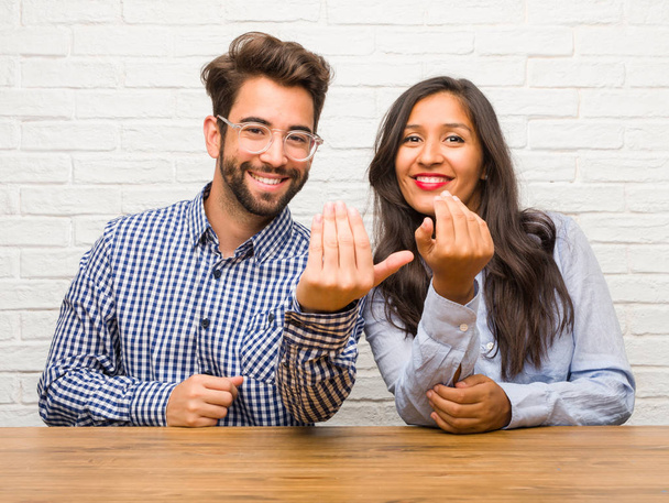 Young indian woman and caucasian man couple inviting to come, confident and smiling making a gesture with hand, being positive and friendly - Photo, Image