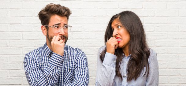 Young indian woman and caucasian man couple biting nails, nervous and very anxious and scared for the future, feels panic and stress - Photo, Image