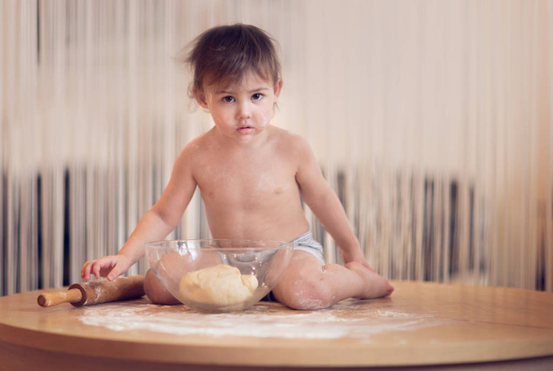 cute boy in flour sitting on the table with raw dough a bowl and looking at the camera  - Фото, изображение