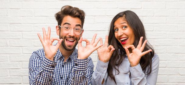 Young indian woman and caucasian man couple cheerful and confident doing ok gesture, excited and screaming, concept of approval and success - Photo, Image