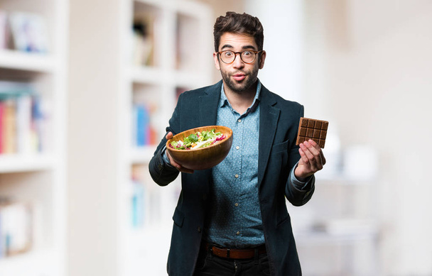 Junger Mann wählt vor verschwommenem Hintergrund zwischen Schokolade und Salat - Foto, Bild