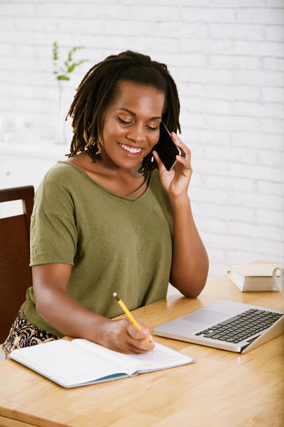 Mulher freelancer sorridente falando ao telefone e tomando notas, escrevendo em diário
  - Foto, Imagem