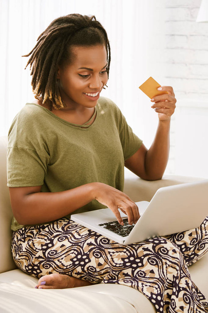 afrikkalais-amerikkalainen iloinen nainen käyttää kannettavaa tietokonetta ja istuu sohvalla ja pitää luottokorttia
  - Valokuva, kuva