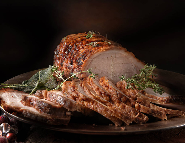 A perfectly oven roasted prime pork joint carved and ready to serve with thyme and sage herbs. Shot against a dark rustic background with generous accommodation for copy space. - Photo, Image