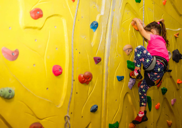 little girl in a pink T-shirt climbing a rock wall indoor - Фото, изображение
