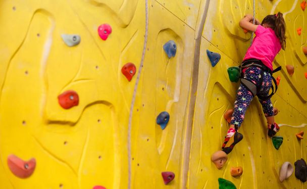 little girl in a pink T-shirt climbing a rock wall indoor - Photo, image