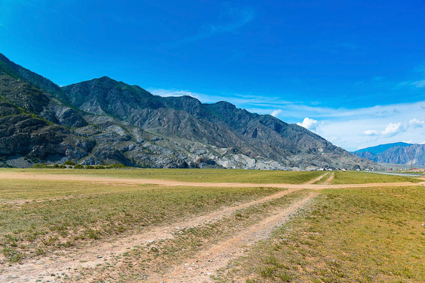 Paysage montagneux de la République d'Altaï, Russie. Un champ avec un chemin de terre au premier plan, de grandes montagnes, loin un village et une petite partie d'une route asphaltée
 - Photo, image