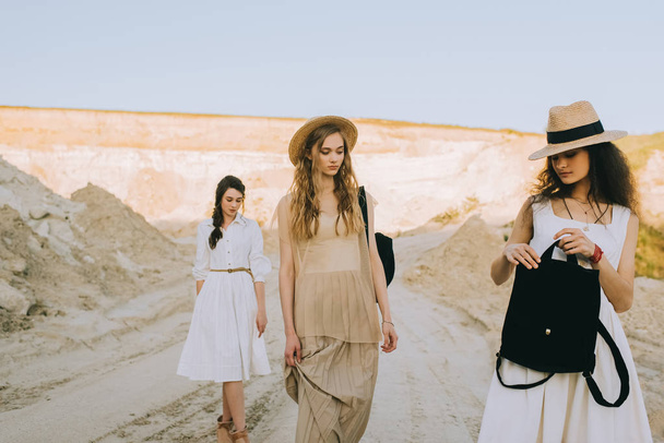 beautiful stylish girlfriends in straw hats walking with backpacks in sandy canyon - Photo, Image