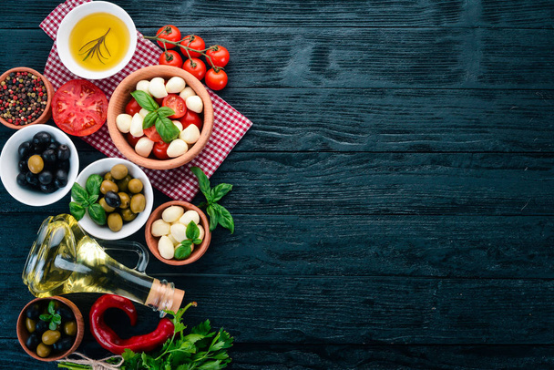 Ingredients for Italian caprese salad. Mozzarella cheese, cherry tomatoes, basil leaves, olives, oil, pepper. On a black wooden background. Free space for text. - Foto, Imagem