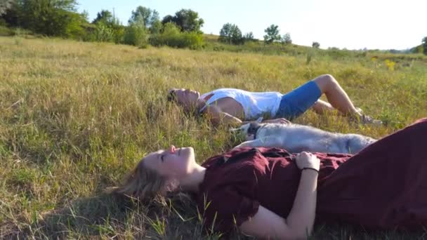 Dolly shot of young couple in love lying on green grass at field and stroking their siberian husky dog at sunny day. Happy pair relaxing and enjoying summer weekend at sunset. Low angle view Close up - Footage, Video