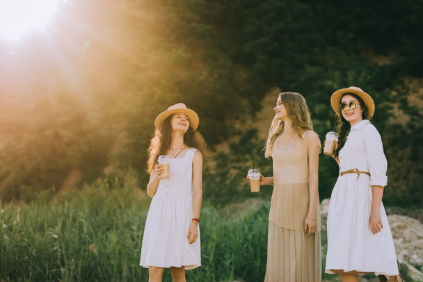 beautiful female friends in straw hats walking in nature with plastic cups of coffee latte - Foto, Bild