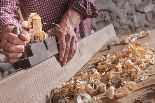 Hombre carpintero raspando trozos de madera rizada con herramienta de mano plana y tablón de madera. Pared de piedra borrosa en el fondo
. - Foto, imagen