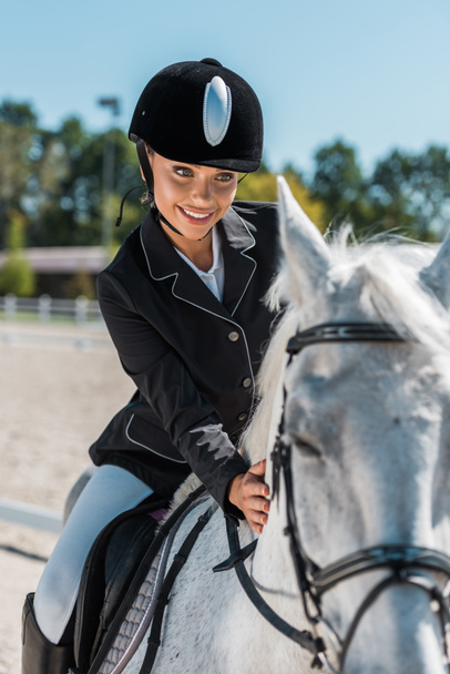 smiling attractive female equestrian in professional apparel riding horse at horse club - Photo, Image