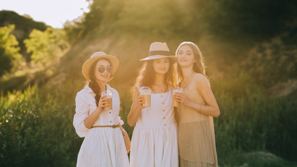 elegantes novias elegantes en sombreros de paja sosteniendo tazas con café con leche y posando, con luz solar
 - Foto, imagen