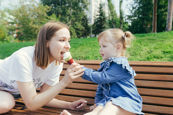 Portret van een jonge moeder en haar dochter van 3 jaar oud eten van ijs in een stadspark op een warme zonnige dag - Foto, afbeelding