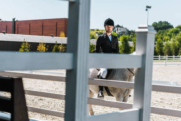 view through fence on handsome male equestrian riding horse at horse club and looking at camera - Photo, Image