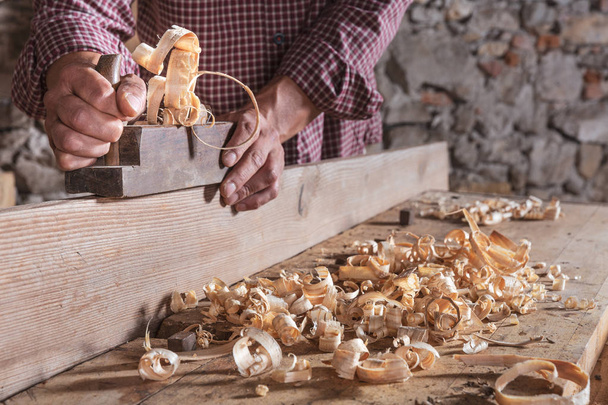 Ο άνθρωπος ξυλουργός απόξεση κατσαρά ξύλινων απορριμάτων με εργαλείο χειρός αεροπλάνο και ξύλινη σανίδα. Θολές πέτρινο τοίχο στο φόντο. - Φωτογραφία, εικόνα